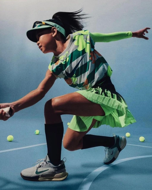 Female tennis player in action with a green and white outfit on an indoor court with scattered tennis balls, sporting a visor and athletic shoes.