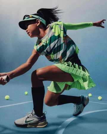 Female tennis player in action with a green and white outfit on an indoor court with scattered tennis balls, sporting a visor and athletic shoes.