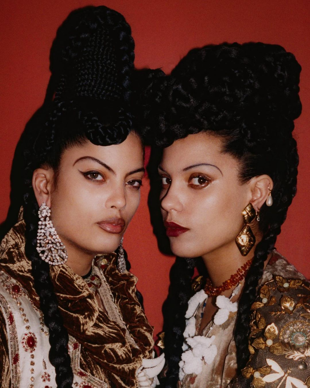 Two women with elaborate braided hairstyles pose in front of a red background wearing embellished tops and statement earrings.