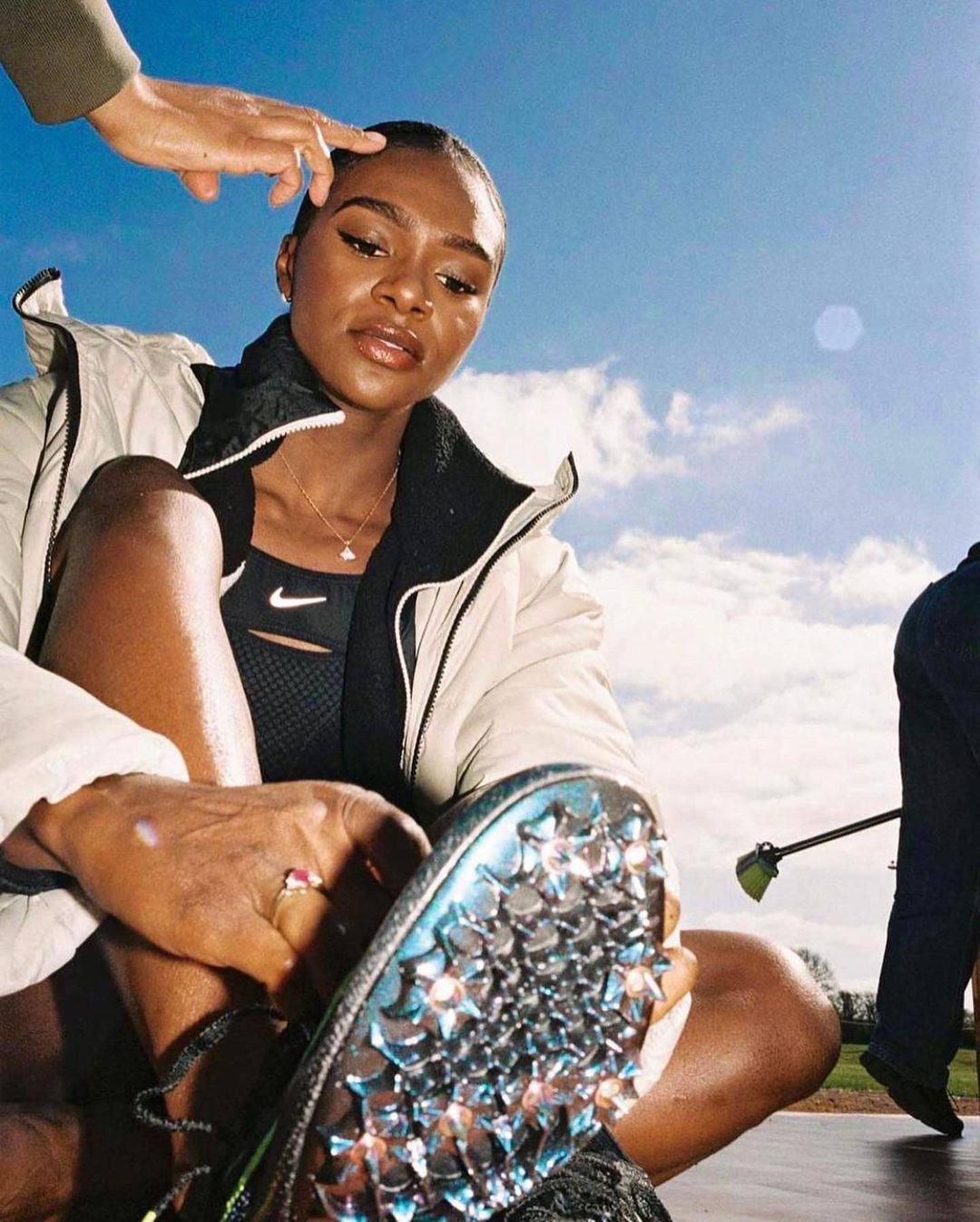 Female athlete in sportswear adjusts her spiked running shoe on a track under a blue sky with clouds; hand hovers above head.