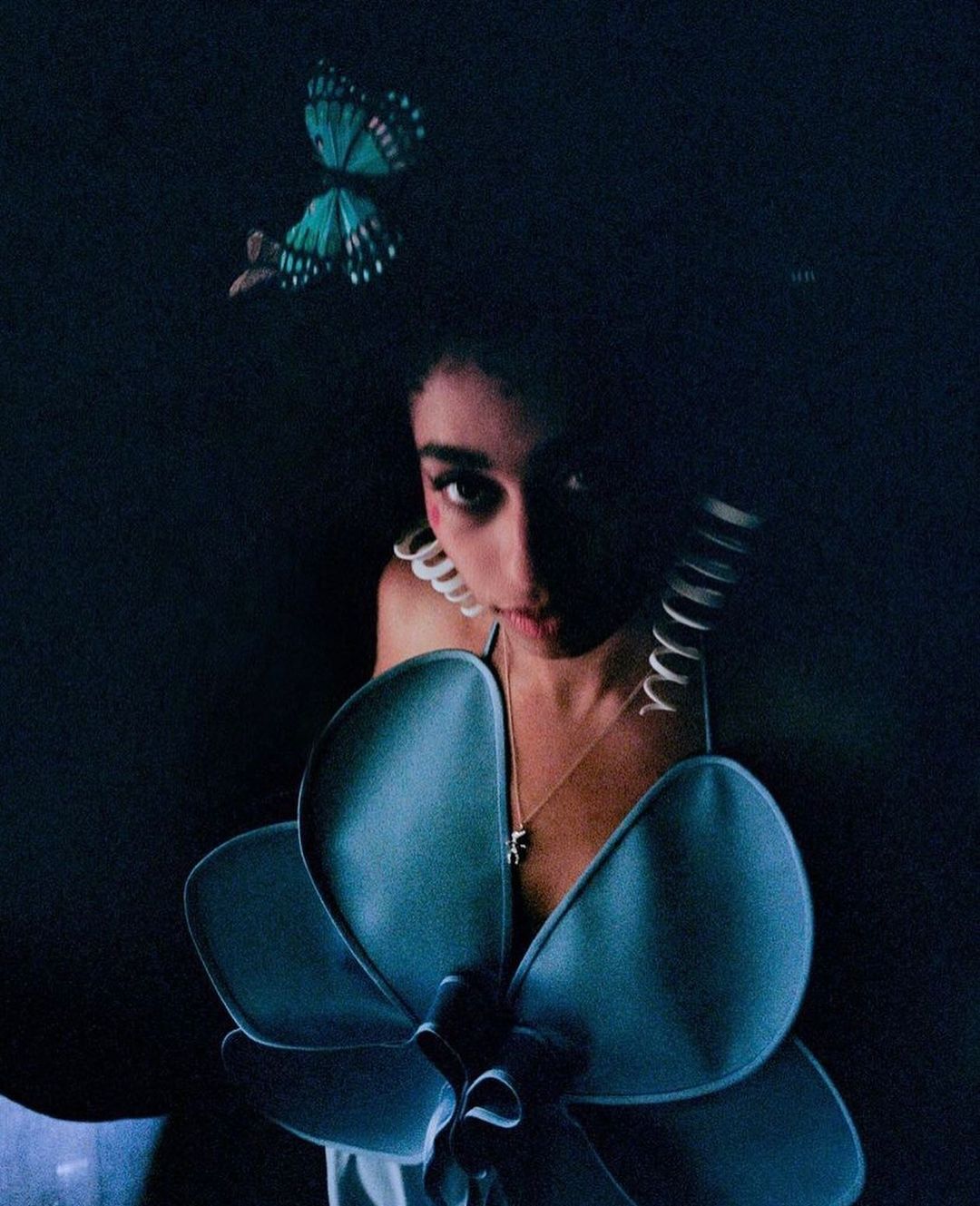 Woman in dark setting wearing a blue floral top and butterfly hair accessory, staring at the camera with a neutral expression.