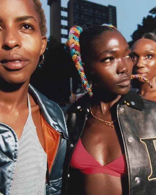 Three Black women stand outdoors, styled in brightly-colored hair accessories and fashionable jackets against an urban backdrop.