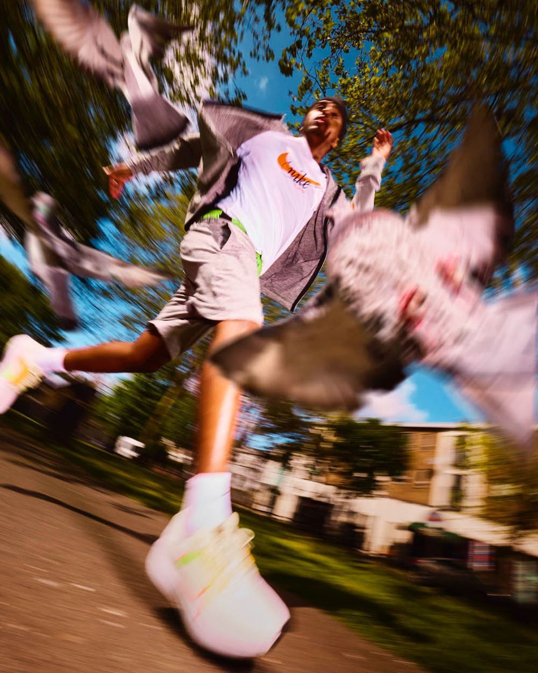 An adult runs forward surrounded by flying pigeons in blurred motion, wearing sportswear, on a paved ground with buildings and trees.