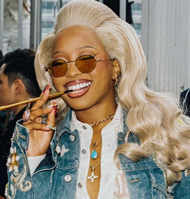 Woman with statement platinum wig and golden glasses smiling, wearing a denim jacket with an ornate cigarette holder in her mouth.