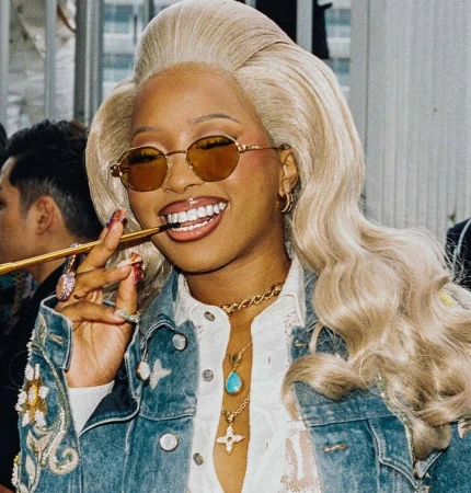 Woman with statement platinum wig and golden glasses smiling, wearing a denim jacket with an ornate cigarette holder in her mouth.