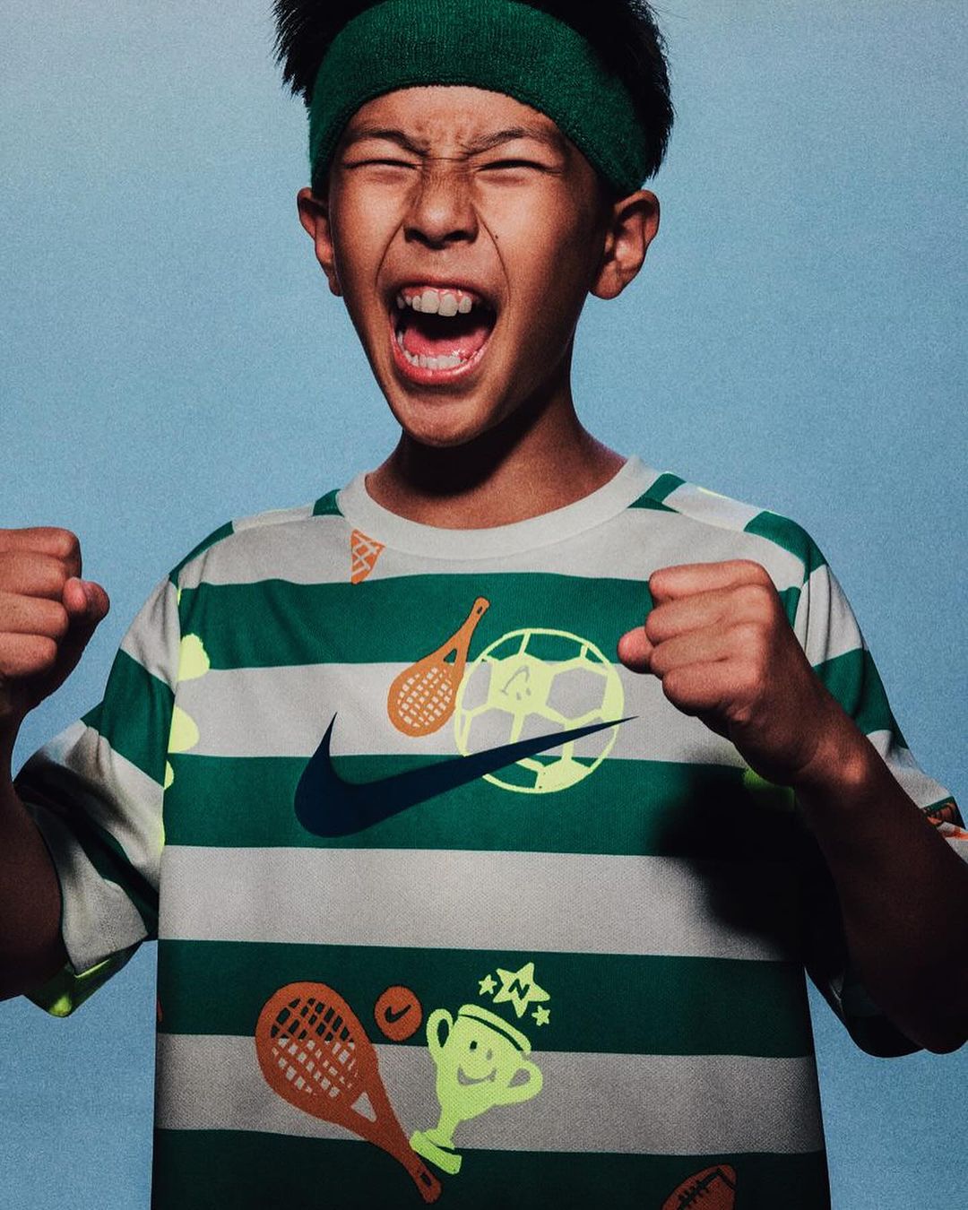 Enthusiastic young boy with a headband wearing a striped shirt with soccer and tennis ball graphics smiles with his fists clenched.