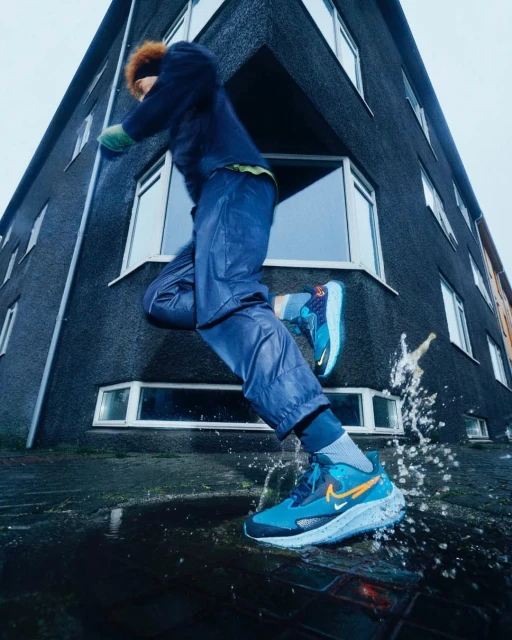 Person in motion wearing blue and teal Nike running shoes, jumping over a puddle near a dark building in an urban outdoor environment.