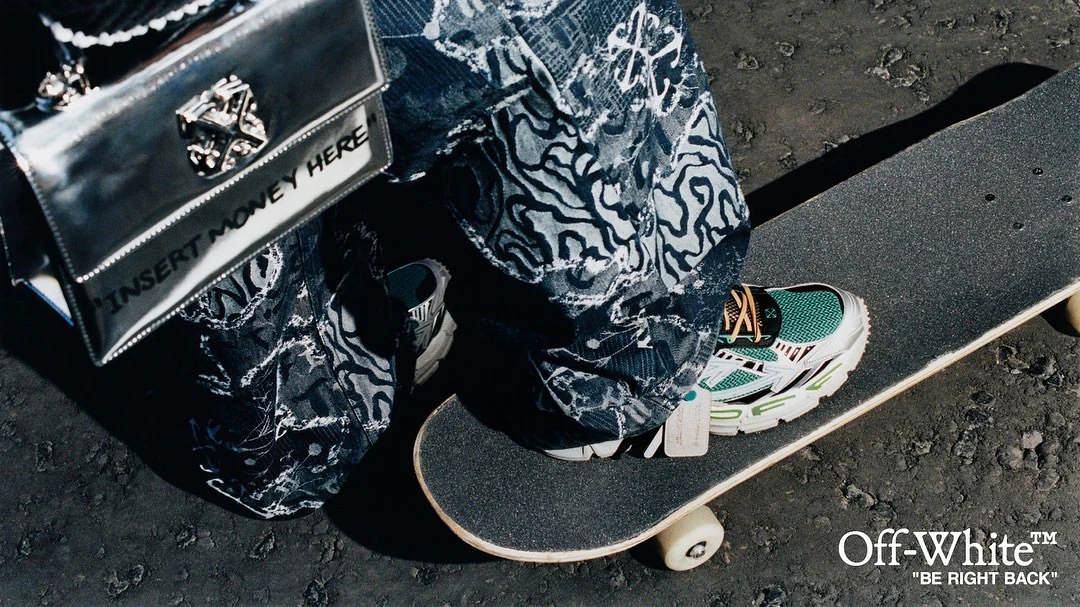 Fashion shot of person on skateboard wearing patterned jeans, sneakers, and a silver purse that says, 'Insert Money Here'.