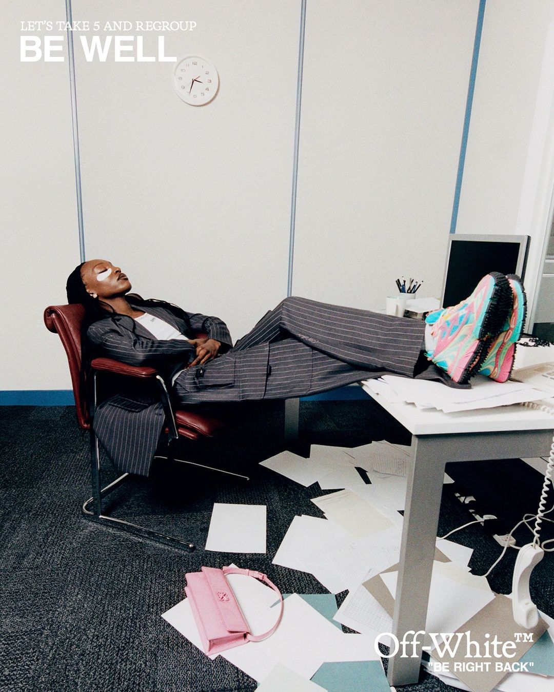 Adult woman wearing a pinstripe suit and colorful sneakers reclining in an office chair with her feet on a cluttered desk.
