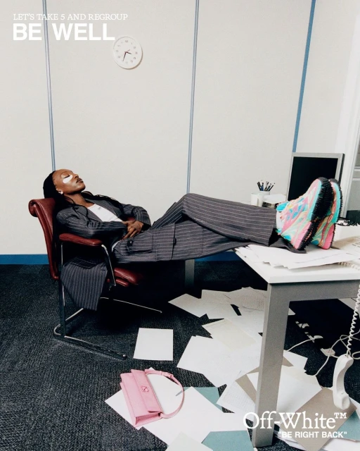 Adult woman wearing a pinstripe suit and colorful sneakers reclining in an office chair with her feet on a cluttered desk.