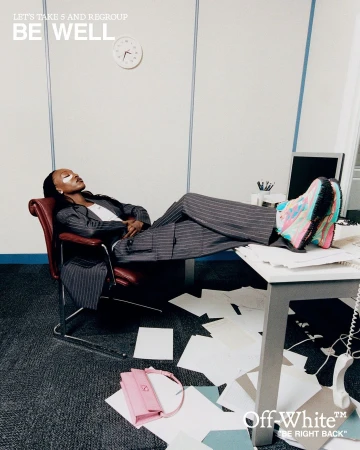 Adult woman wearing a pinstripe suit and colorful sneakers reclining in an office chair with her feet on a cluttered desk.