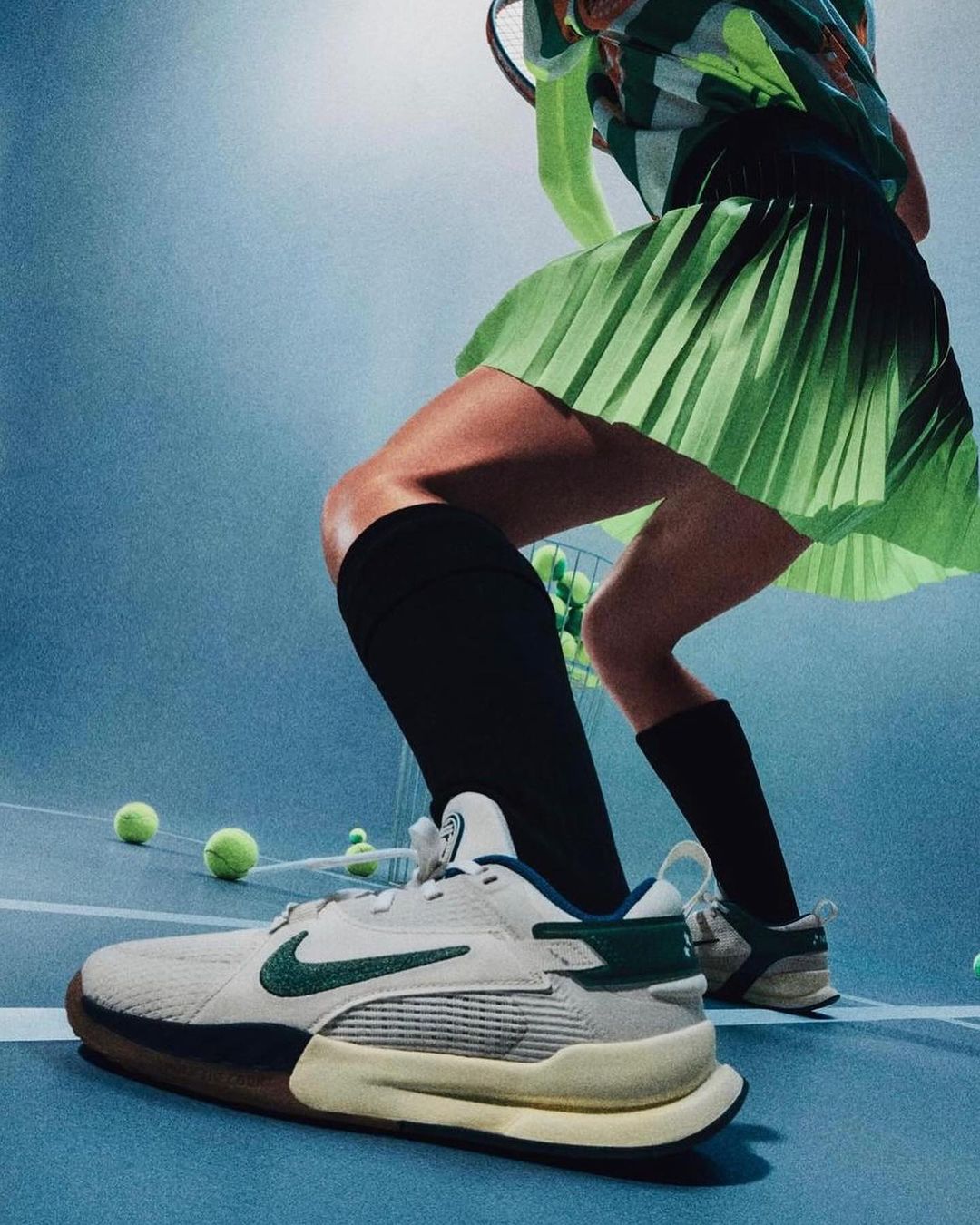 Close-up of tennis shoe on blue court with tennis balls, tennis player in blurred background wearing green skirt and holding racket.
