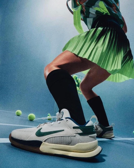Close-up of tennis shoe on blue court with tennis balls, tennis player in blurred background wearing green skirt and holding racket.