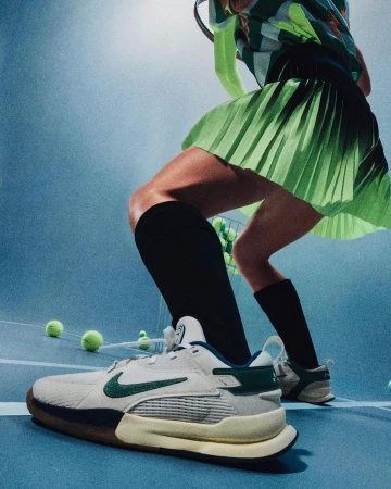 Close-up of tennis shoe on blue court with tennis balls, tennis player in blurred background wearing green skirt and holding racket.