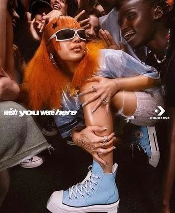 Woman with orange hair wearing sunglasses and blue Converse sneakers in a fashion shot surrounded by other people in an indoor setting.