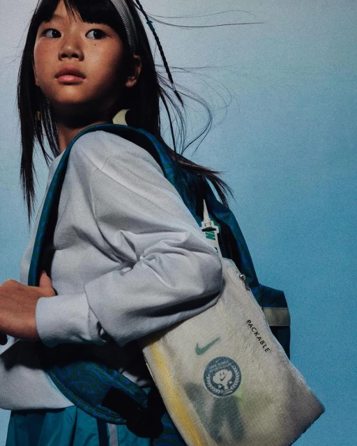Asian child with backpack posing in front of a blue backdrop, wearing a white top and blue skirt, showcasing Nike bag with logo.