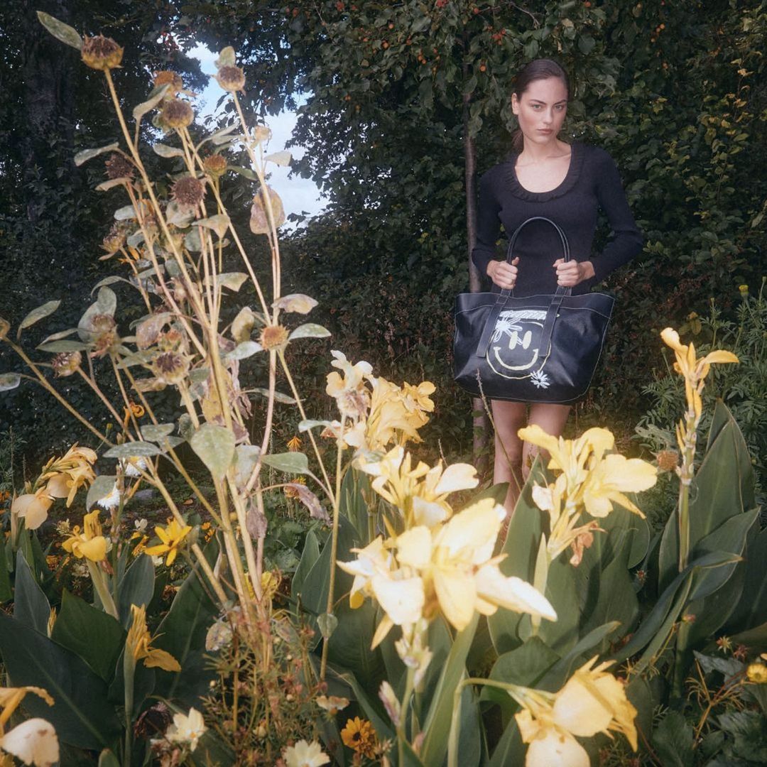 Woman in black dress holds a tote bag, standing among yellow flowers and greenery in a garden setting during daytime.