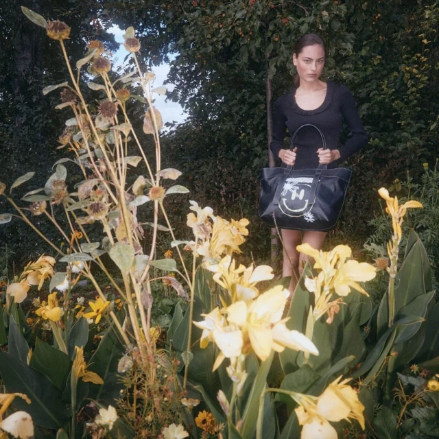 Woman in black dress holds a tote bag, standing among yellow flowers and greenery in a garden setting during daytime.