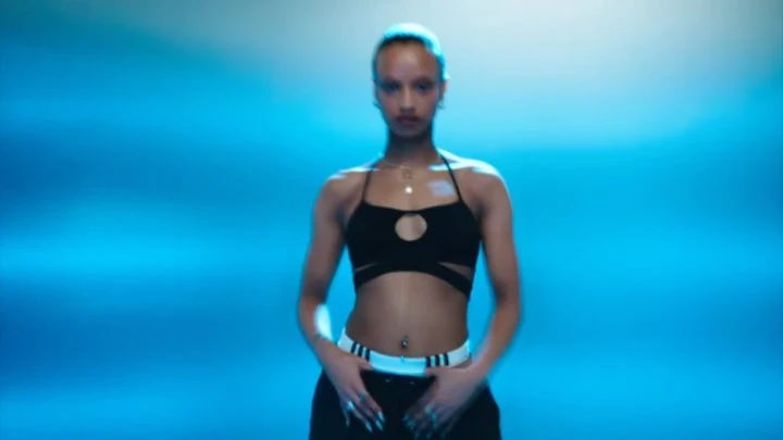 An adult female model stands centered in a brightly lit photo studio wearing black athletic apparel against a vivid blue background.