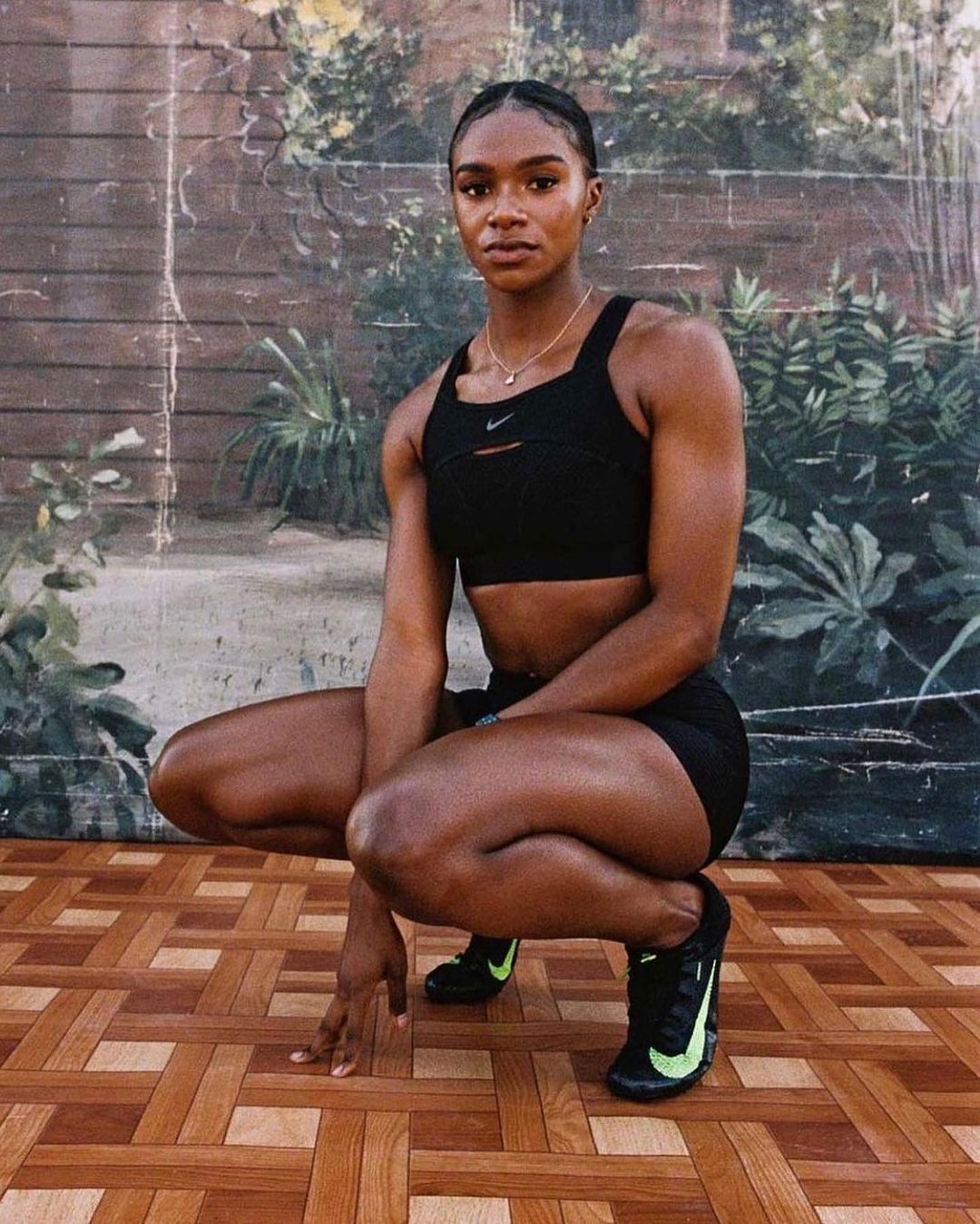Fit African-American woman in black activewear crouches on a parquet floor in front of a hand-painted plant and wall mural indoors.