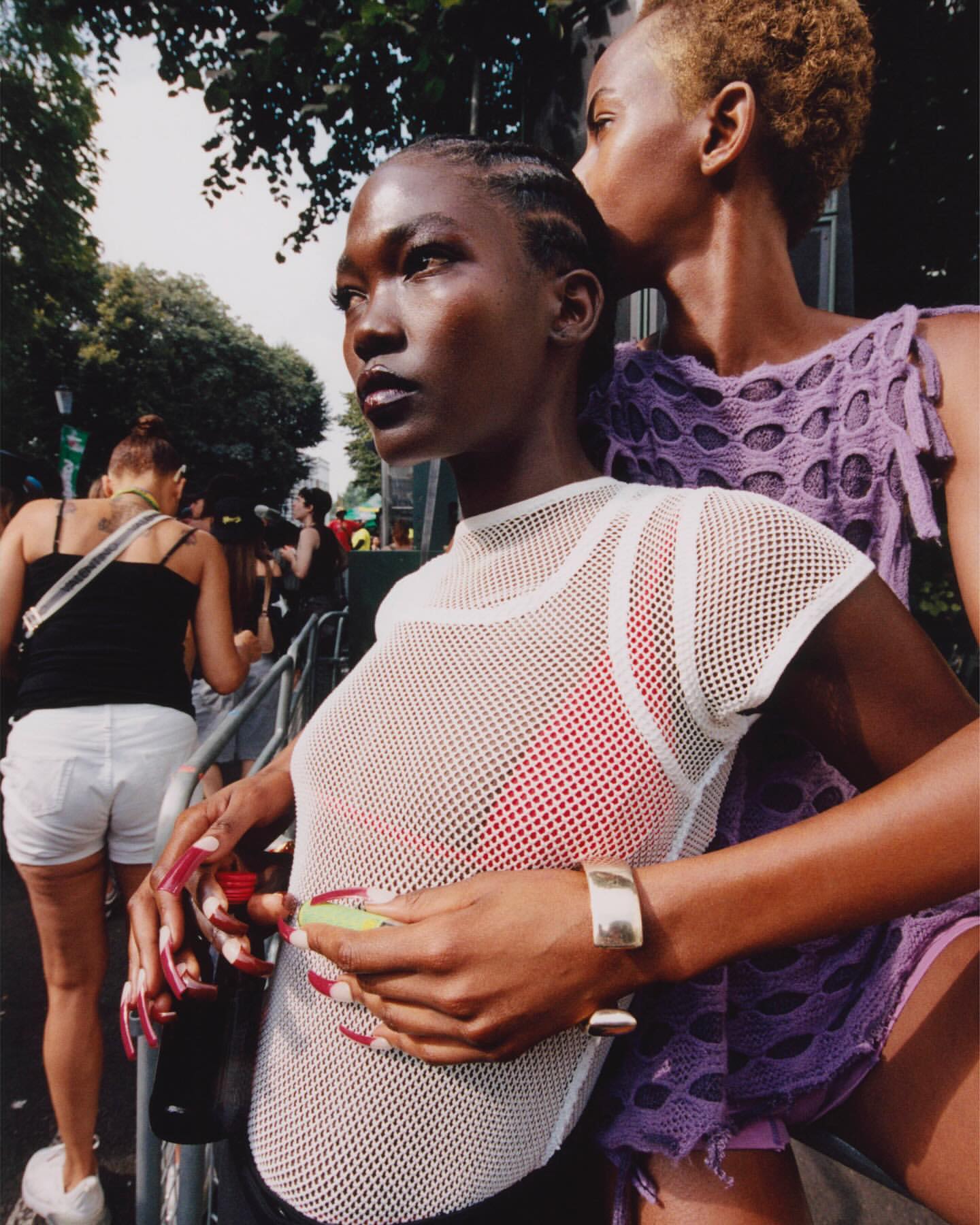 Two women with dark skin pose outdoors. One wears a white mesh top with a red top underneath and the other has a purple crocheted shirt.