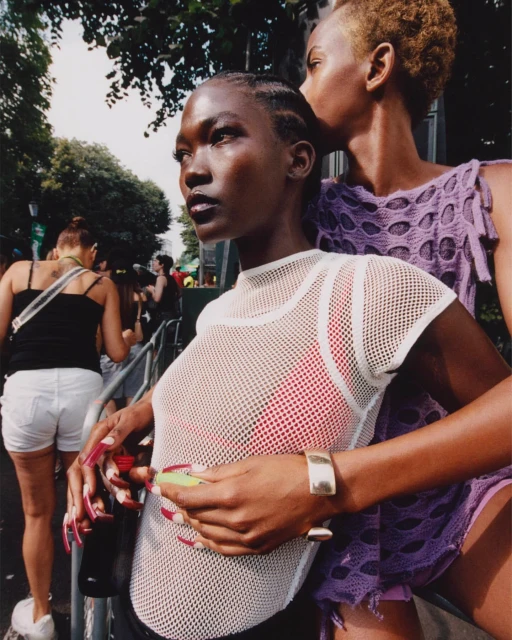Two women with dark skin pose outdoors. One wears a white mesh top with a red top underneath and the other has a purple crocheted shirt.