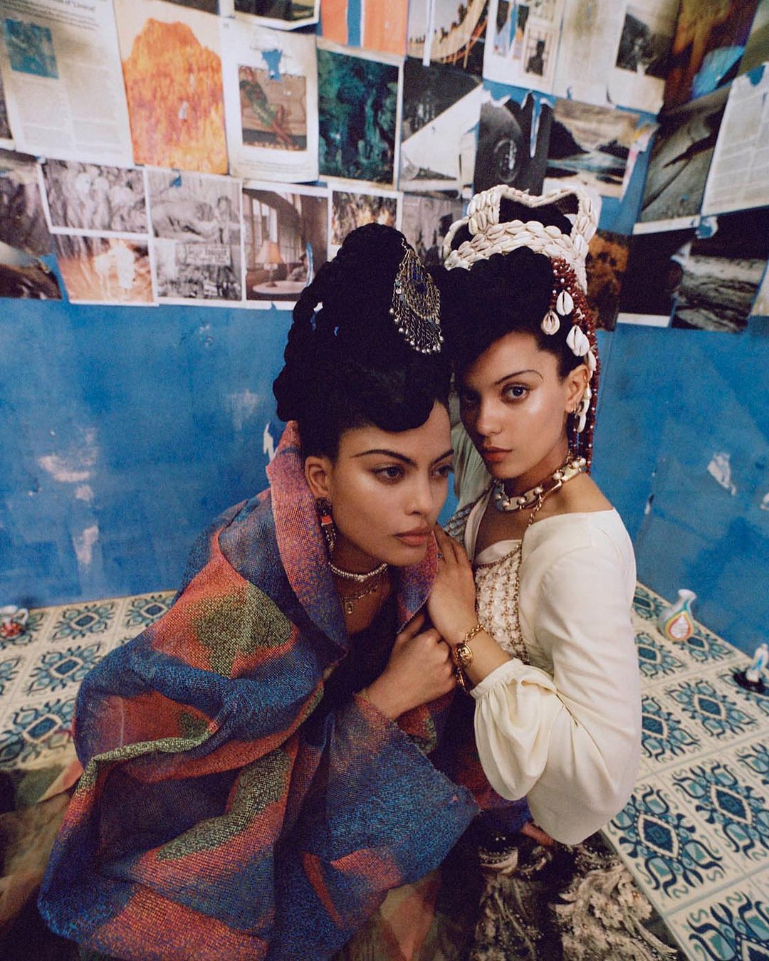Two women in elaborate headwear pose indoors. One wears a colorful wrap; the other, a white blouse and beaded jewelry, with art on the walls.