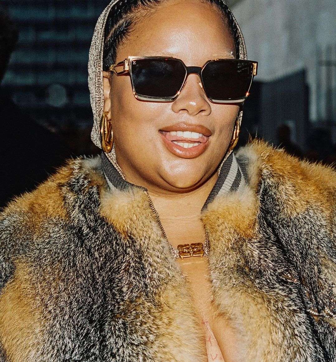 Headshot of an adult woman wearing sunglasses, a fur coat, a head covering, and gold hoop earrings smiling with teeth showing.
