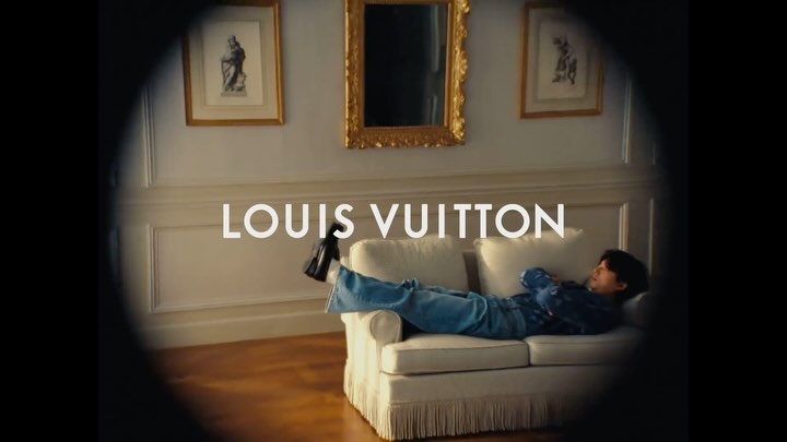 Indoor portrait of a person relaxing on a white couch, wearing blue jeans and a blue patterned shirt with a Louis Vuitton logo.