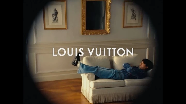 Indoor portrait of a person relaxing on a white couch, wearing blue jeans and a blue patterned shirt with a Louis Vuitton logo.