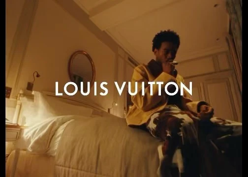 An adult male smoking on a bed inside a bedroom with a gold framed oval mirror, white walls, and a white bed with the text LOUIS VUITTON.
