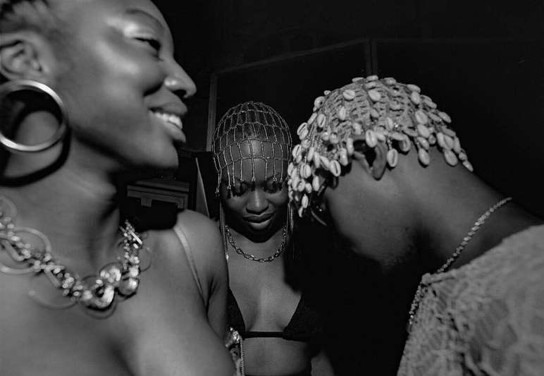 Three Black women are close together in a photo studio, adorned with jewelry and shell-covered hairpieces, captured in monochrome.