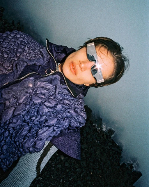 Adult in a puffer jacket and futuristic sunglasses lies on a rocky terrain, posing in an outdoor setting with an overcast sky.