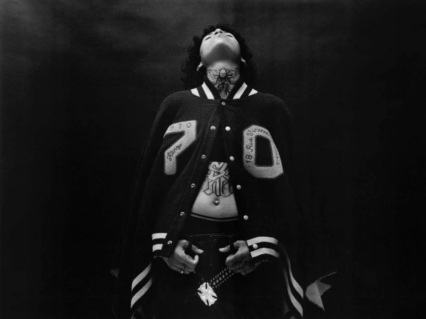A person with tattoos poses in a letterman jacket with '970' on it, against a dark background in a medium studio shot.