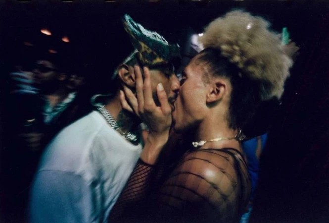 Close-up of two adults kissing in a dimly lit setting, one wearing a white t-shirt and patterned hat, the other in black mesh.
