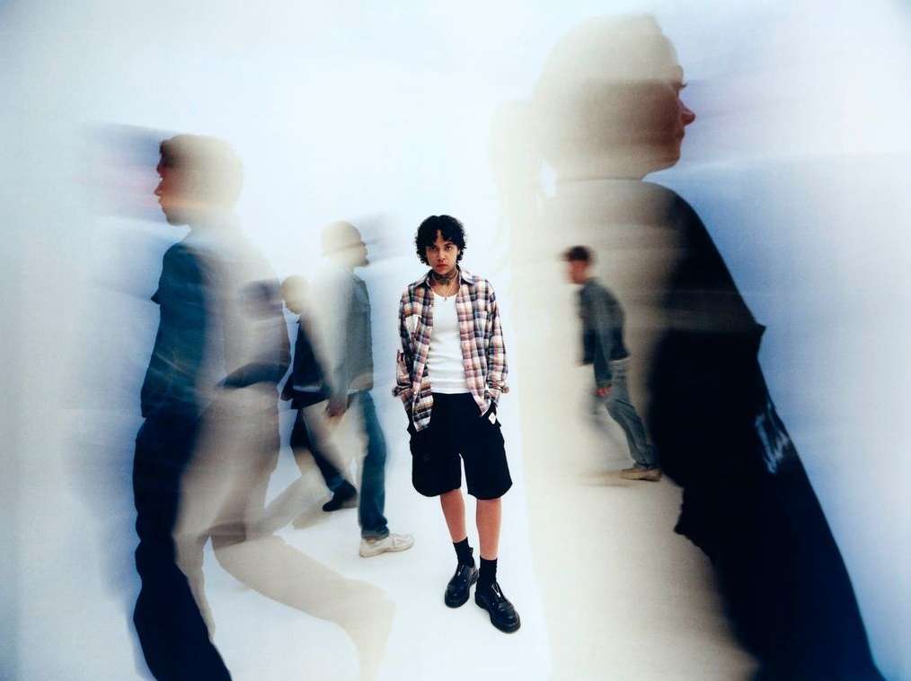 Portrait of a young adult in a plaid shirt standing in a white space, with blurred figures in the foreground and background.