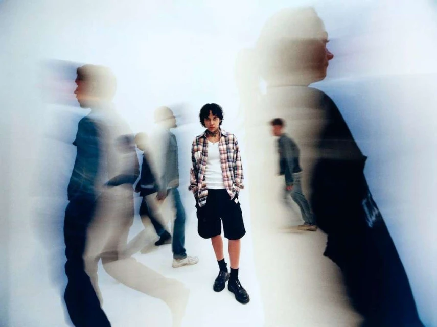 Portrait of a young adult in a plaid shirt standing in a white space, with blurred figures in the foreground and background.