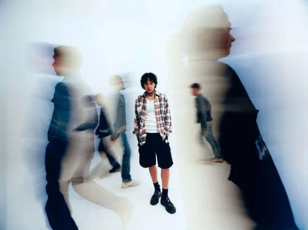 Portrait of a young adult in a plaid shirt standing in a white space, with blurred figures in the foreground and background.