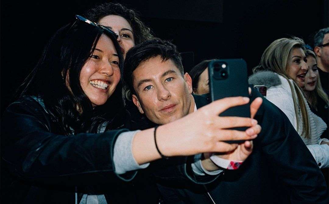 Actor Barry Keoghan poses for a selfie with a smiling Asian woman in an indoor venue, surrounded by a small group of people.