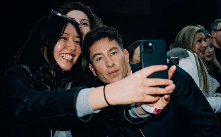 Actor Barry Keoghan poses for a selfie with a smiling Asian woman in an indoor venue, surrounded by a small group of people.