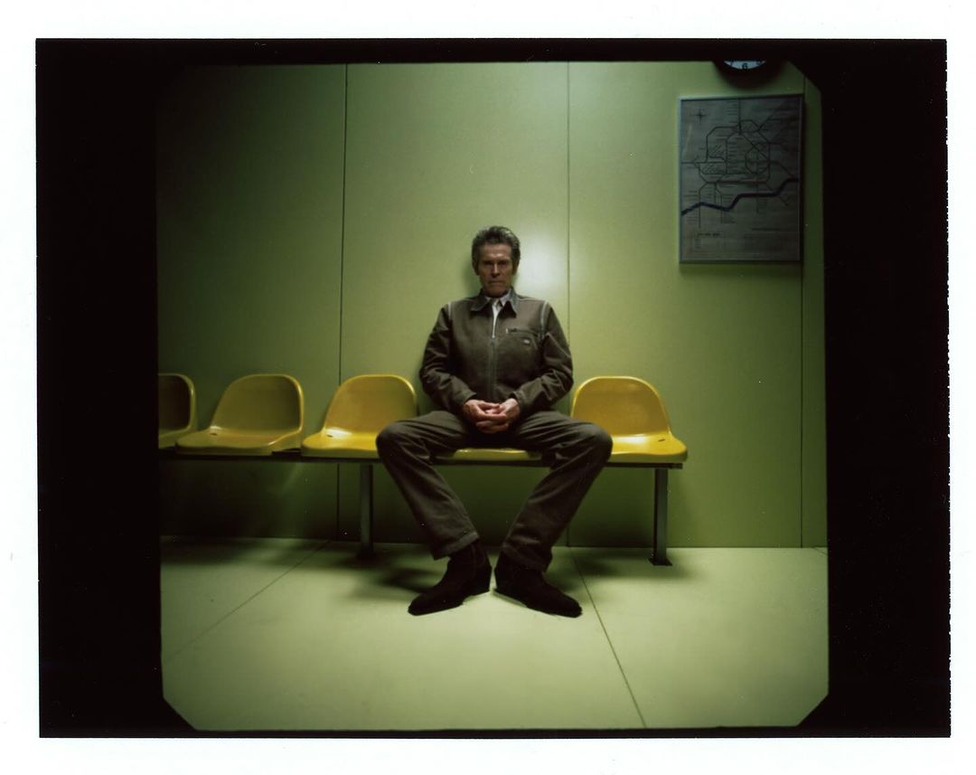 Man in utilitarian jumpsuit seated in waiting room with yellow chairs, in a muted green interior, with a schematic wall map.