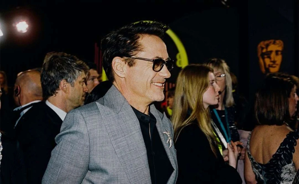 Smiling man in a grey suit jacket and sunglasses at an indoor event surrounded by people, captured in a medium shot.