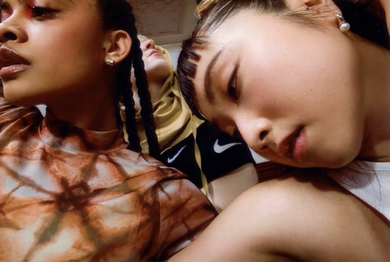 Close-up of three young women with striking makeup, diverse hairstyles, and stylish tops; one wearing a yellow hoodie with the Nike logo.