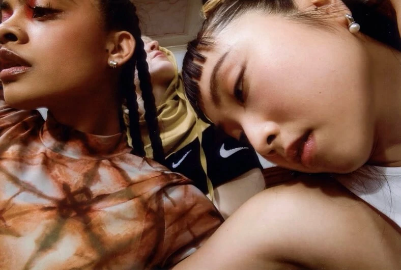 Close-up of three young women with striking makeup, diverse hairstyles, and stylish tops; one wearing a yellow hoodie with the Nike logo.