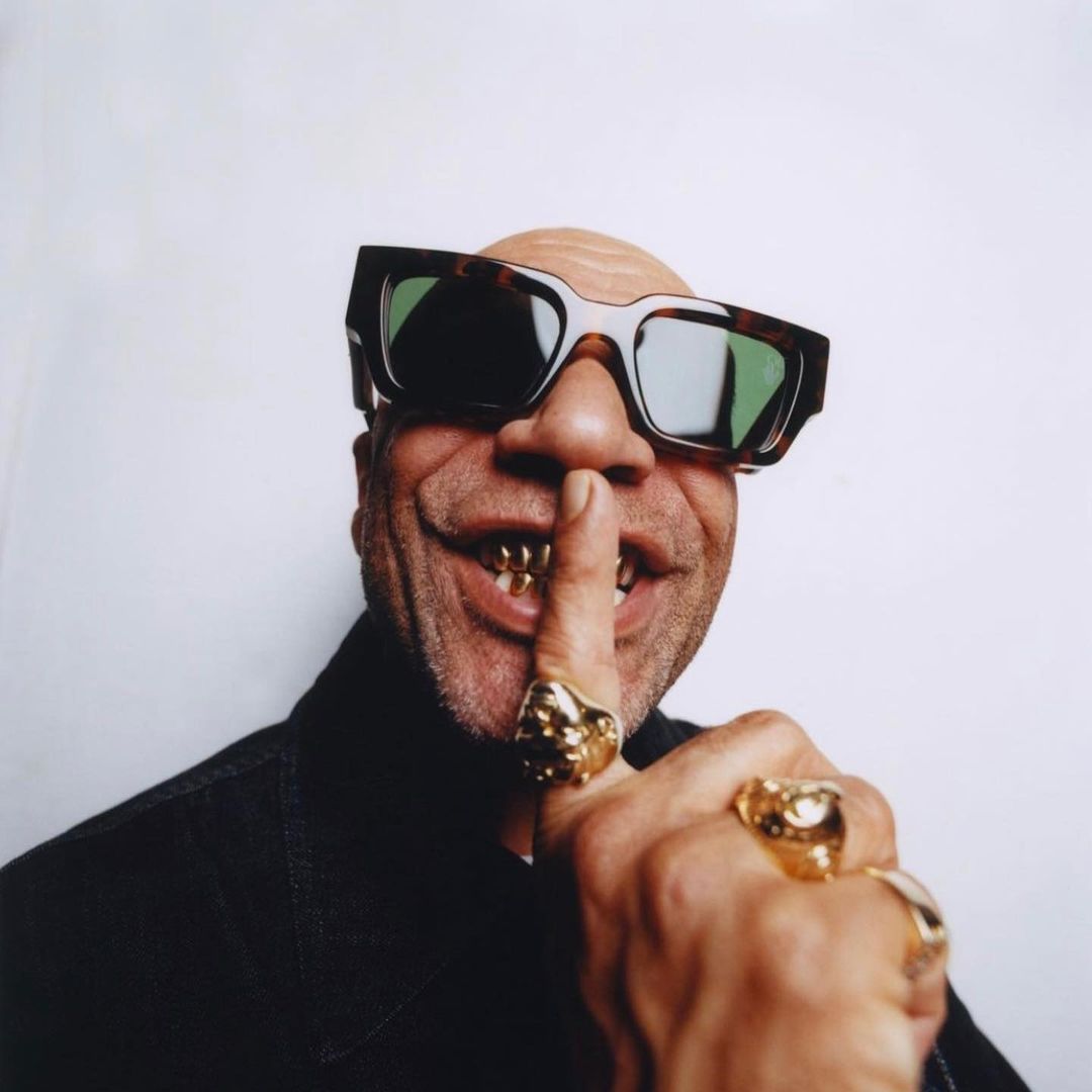Close-up portrait of a bald adult male wearing sunglasses and gold teeth grills, with a finger to his lips, indicating silence.