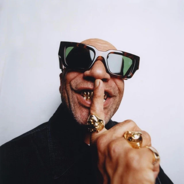 Close-up portrait of a bald adult male wearing sunglasses and gold teeth grills, with a finger to his lips, indicating silence.