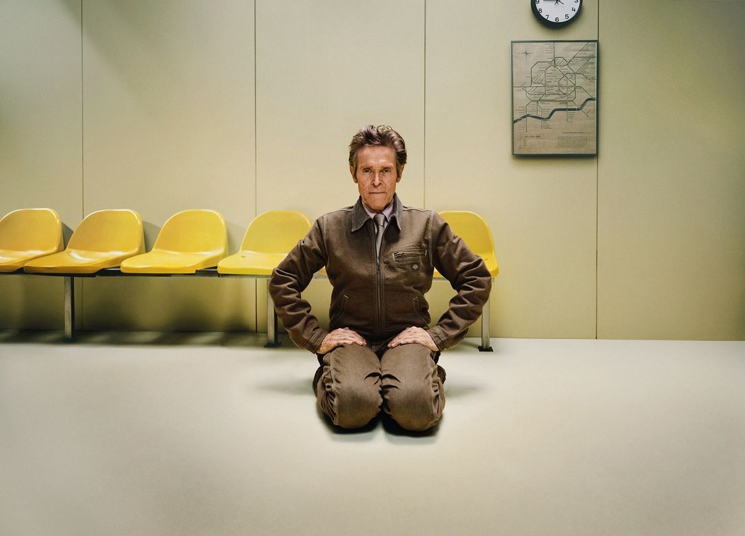 Eye-level shot of an adult man in a brown jumpsuit kneeling indoors, posing against a pale wall with yellow chairs and a framed map.