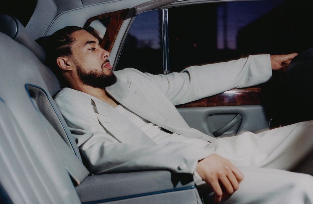Adult male in a white suit relaxes in the back seat of a car, showcasing a profile view with a beard and a calm expression.