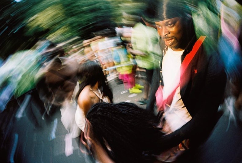 Dynamic crowd scene featuring a Black man in a black jacket and durag, alongside a woman with long dark hair in a blurry street setting.