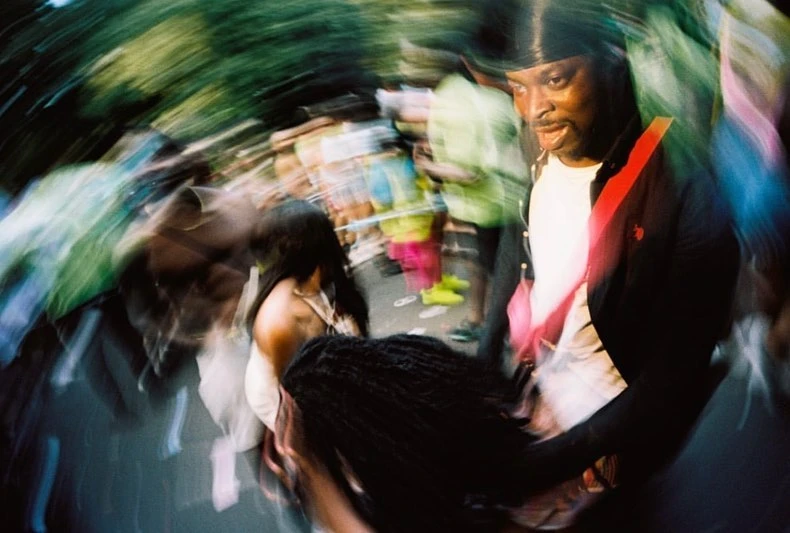 Dynamic crowd scene featuring a Black man in a black jacket and durag, alongside a woman with long dark hair in a blurry street setting.